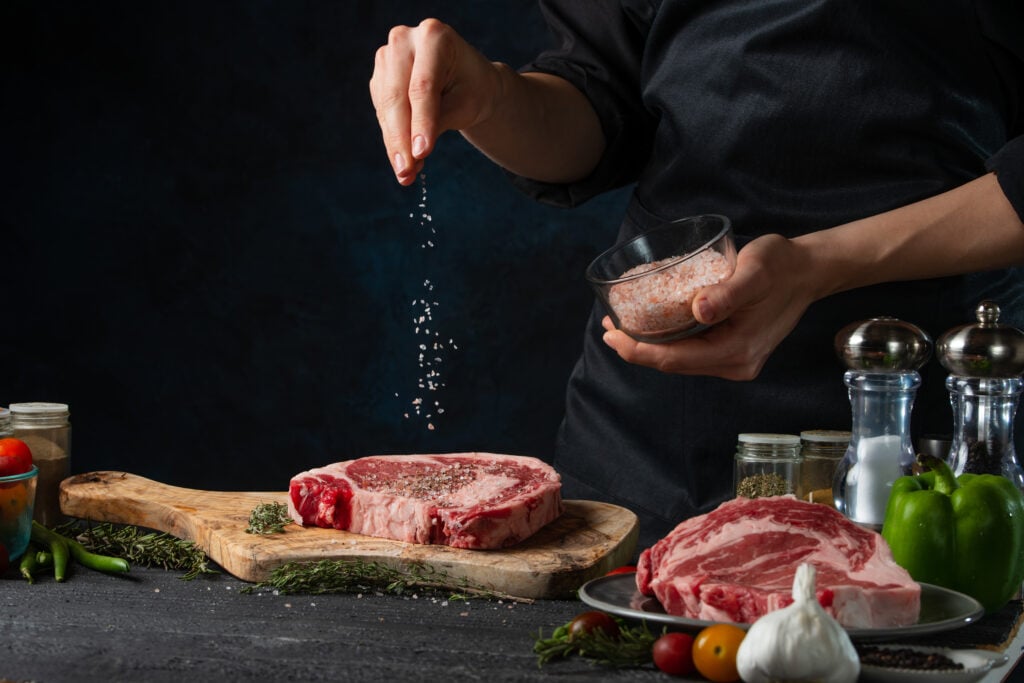 Chef,Pours,Sea,Salt,On,Raw,Steak,On,Wooden,Chopped